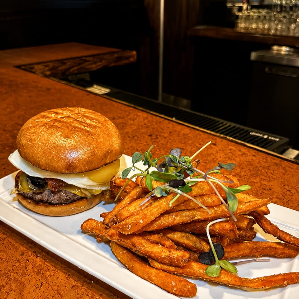 Gourmet burger with a fried egg and sweet potato fries on a white plate at a bar.