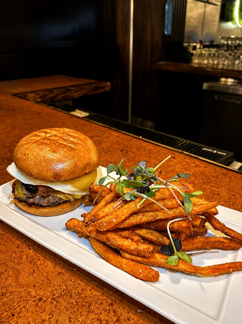 Gourmet burger with a fried egg and sweet potato fries on a white plate at a bar.