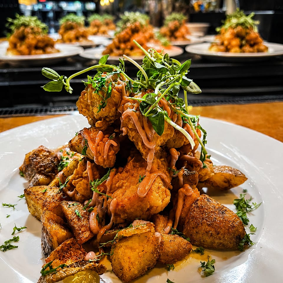 Crispy fried chicken and roasted potatoes topped with sauce and microgreens, with more plates in the background.