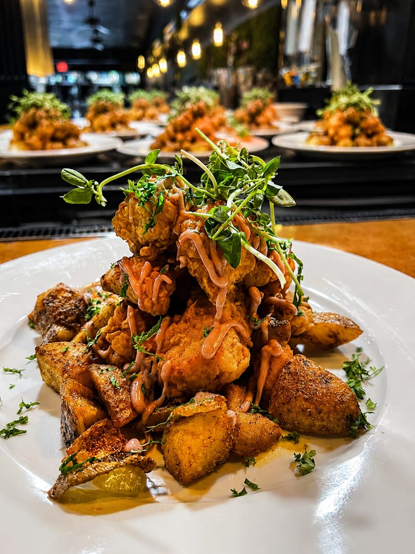 Crispy fried chicken and roasted potatoes topped with sauce and microgreens, with more plates in the background.