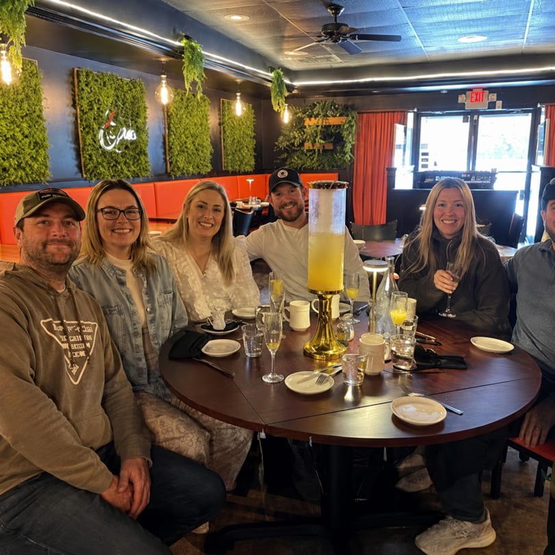 Six smiling people gathered around a round table with drinks in a restaurant with green wall decor.