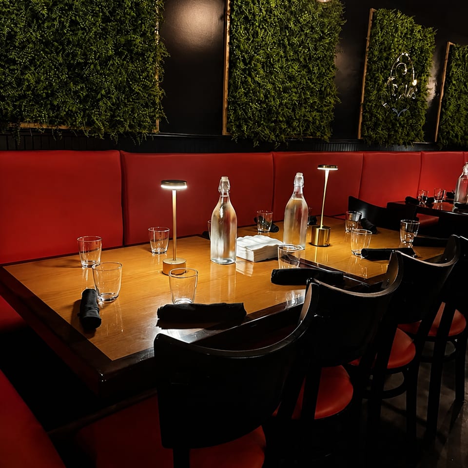 A long dining table with red booth seating, black chairs, and green plant wall decor in a restaurant.