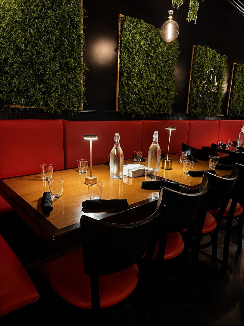 A long dining table with red booth seating, black chairs, and green plant wall decor in a restaurant.