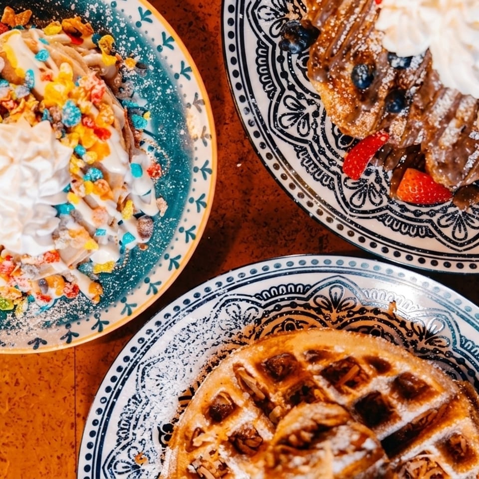 Three colorful waffles on decorative plates, topped with fruit, cereal, whipped cream, and powdered sugar.