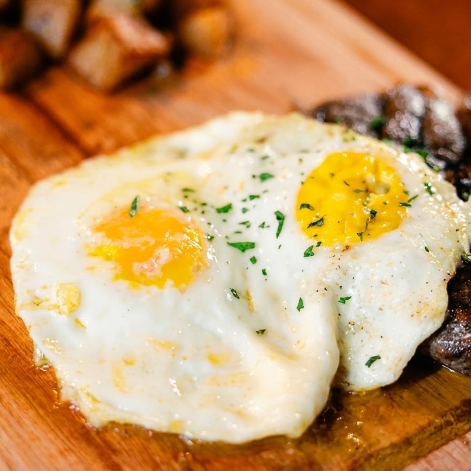 Two fried eggs with parsley, steak, and roasted potatoes on a wooden board.