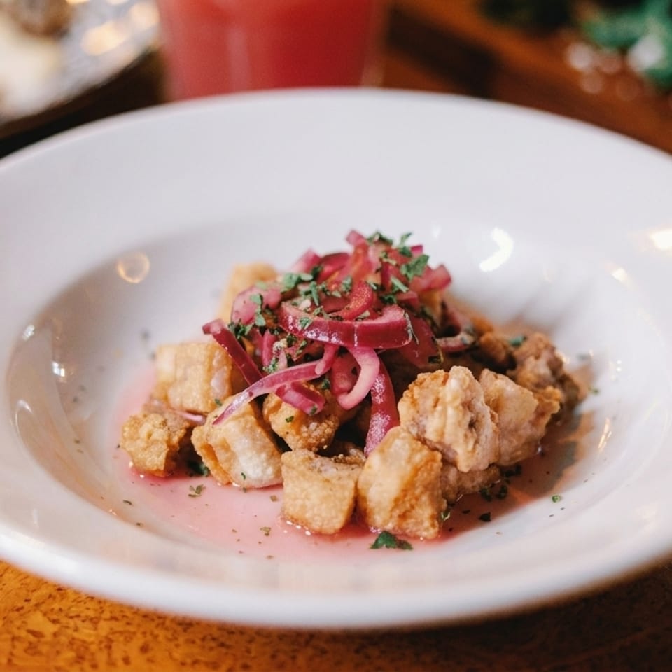 Crispy fried pork belly pieces topped with pickled red onions and herbs in a white bowl.